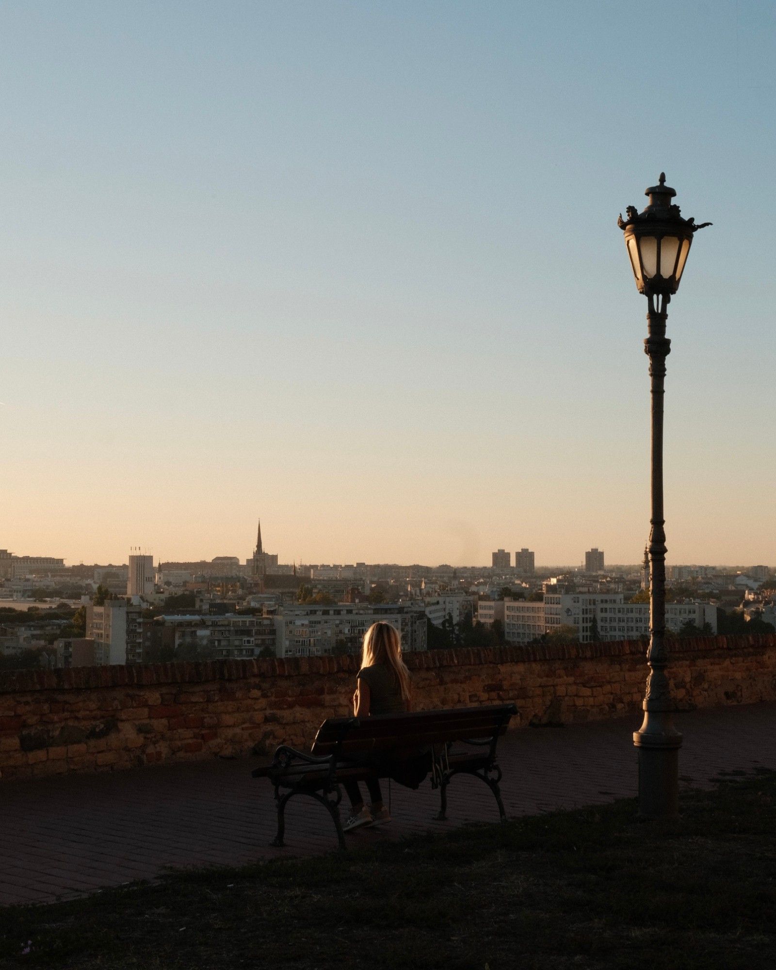 A woman on a bench next to a street lamp, city skyline at sunset